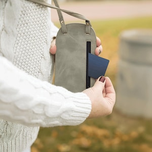 May include: A gray, leather-like crossbody phone case with a strap. The case has a vertical zippered pocket and a dark blue card partially inserted. The case is being held by a person wearing a white cable-knit sweater.