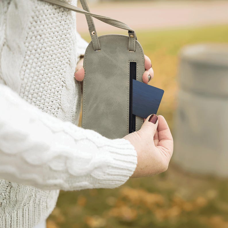 May include: A gray, leather-like crossbody phone case with a strap. The case has a vertical zippered pocket and a dark blue card partially inserted. The case is being held by a person wearing a white cable-knit sweater.