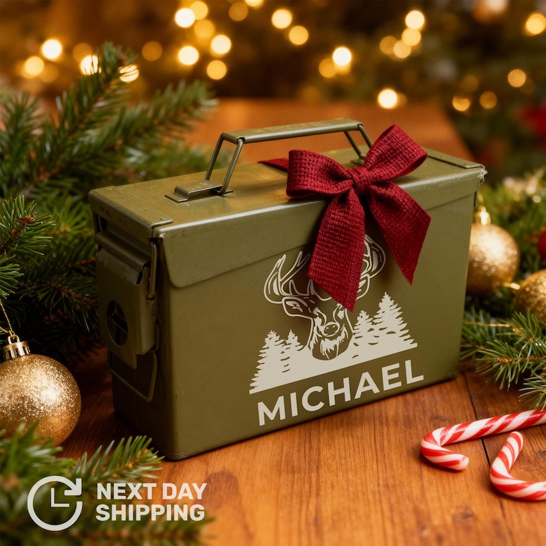 May include: An olive green metal box with a handle, decorated with a white deer and tree design, and the name "MICHAEL". A burgundy bow adorns the top. The box is on a wooden surface, surrounded by Christmas decorations.