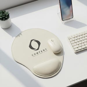 May include: A cream-colored ergonomic mouse pad with a wrist rest, a white wireless mouse, and a white keyboard are on a white desk. The mouse pad has a black logo and the text "COMPANY TAGLINE GOES HERE". A small succulent plant in a white pot and a smartphone are also on the desk.