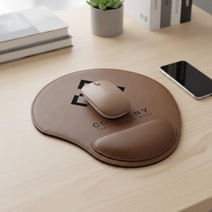 May include: A brown leatherette mouse pad with a built-in wrist rest and a wireless mouse. The mouse pad has a printed logo and text. A smartphone and a stack of books are also on the light wood surface.