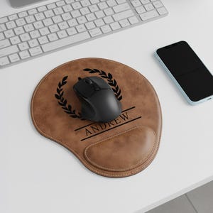 May include: A brown leather mousepad with a black mouse, a black laurel wreath design, and the name "ANDREW" in a black font. A black smartphone and a white keyboard are also visible.
