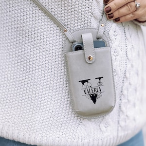 May include: A light gray leather phone crossbody bag with a strap. The bag has a snap closure and a black graphic with the name "Valeria". The phone is visible inside the bag.