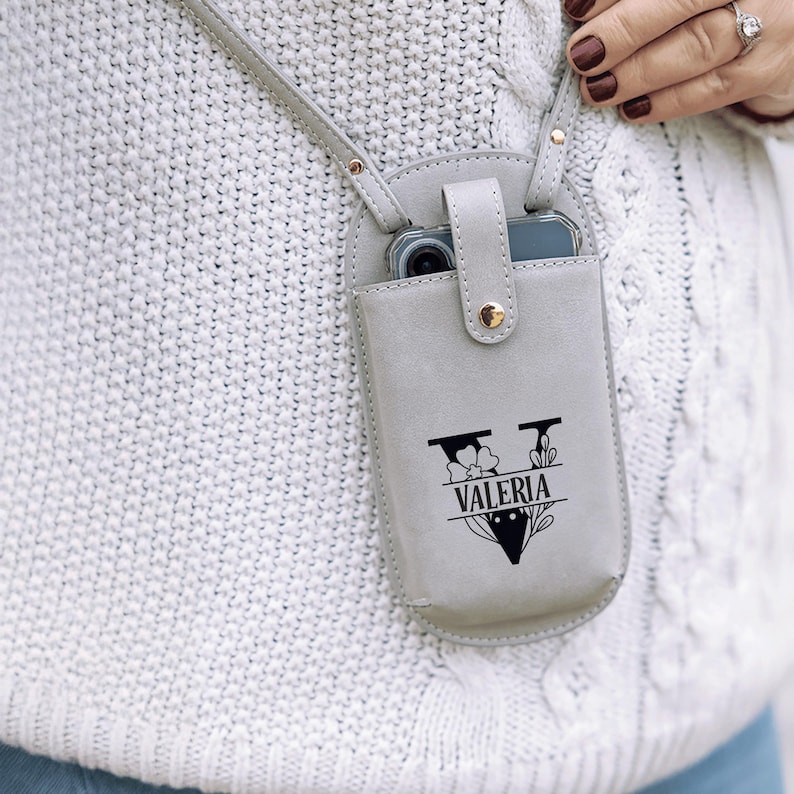 May include: A light gray leather phone crossbody bag with a strap. The bag has a snap closure and a black graphic with the name "Valeria". The phone is visible inside the bag.