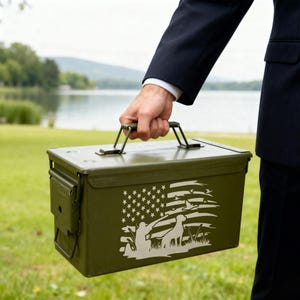 May include: Olive green metal storage box with a handle, featuring a white graphic of the American flag, a hunter, and a dog. The box is being held outdoors near a lake.