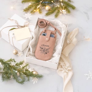 May include: A blush pink leather phone case with a floral design and the name "Isabella" engraved on the front. The case has a matching strap and is presented in a white gift box with tissue paper.