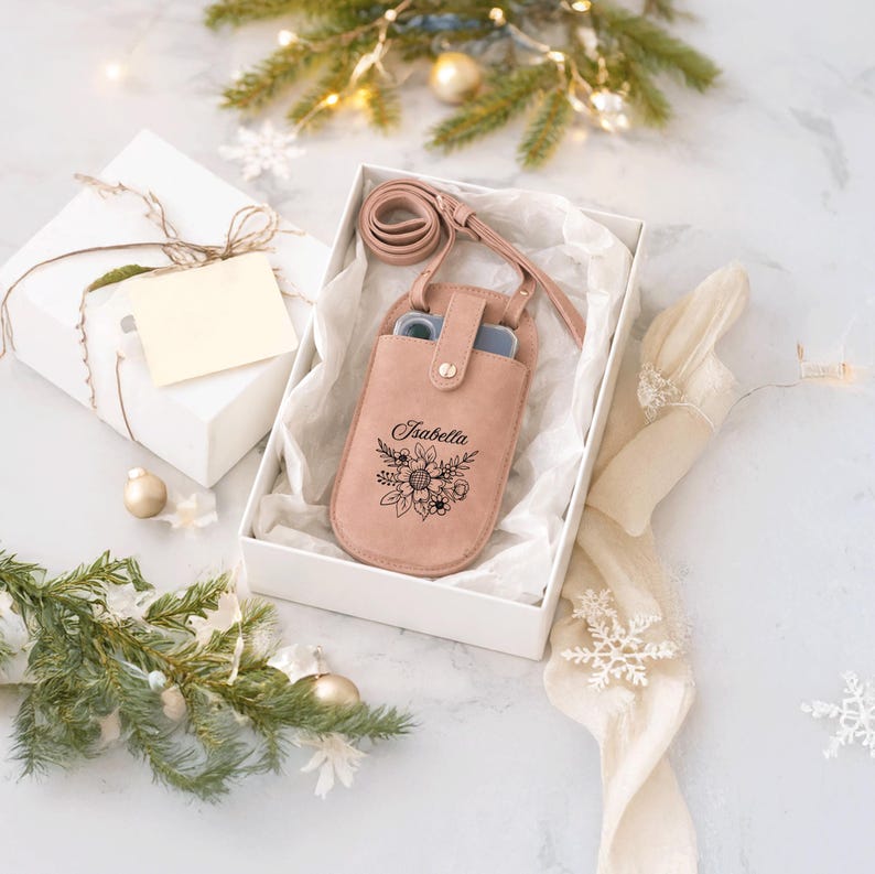 May include: A blush pink leather phone case with a floral design and the name "Isabella" engraved on the front. The case has a matching strap and is presented in a white gift box with tissue paper.