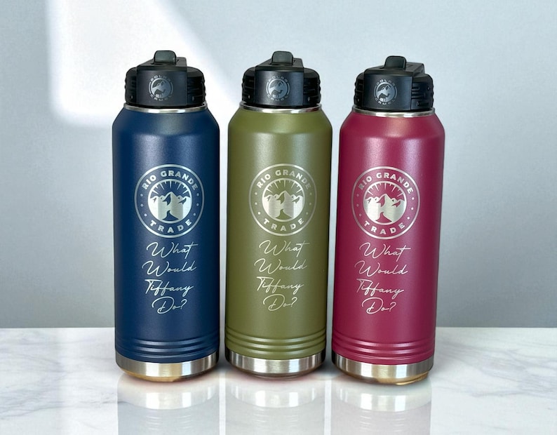 Three insulated water bottles in navy blue, olive green, and maroon stand side by side on a white surface, each engraved with a circular logo reading Rio Grande Trade and the phrase What Would Tiffany Do?