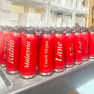 Puede incluir: Una colecci&oacute;n de botellas de agua aisladas de color rojo con tapas negras y bases plateadas. Cada botella tiene un nombre grabado en una fuente blanca y elegante. Las botellas est&aacute;n dispuestas sobre una superficie gris, con estantes y cajas en el fondo.