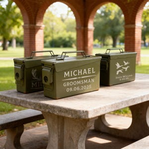 May include: Three olive green metal boxes with handles, sitting on a stone table. The boxes have white text and graphics, including names, dates, and a deer antler design. The boxes are likely for storage or gifts.
