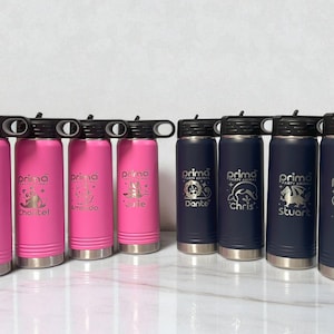 A row of eight insulated water bottles with black lids standing on a white surface, featuring four bright pink bottles on the left and four dark navy blue bottles on the right, all engraved with custom animal graphics and names.