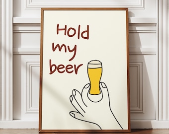 May include: A framed art print with the words "Hold my beer" in brown lettering. The image features a cartoon hand holding a glass of yellow beer with white foam. The background is a cream color.