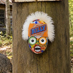 May include: A decorative oval mask with a white faux fur trim, featuring a colorful abstract pattern, large green and white eyes, a wide mouth with teeth, and the name BILLY printed in blue letters on an orange background, mounted on a wooden post.