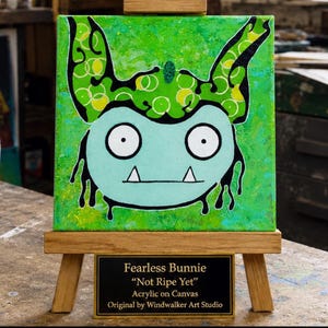 Puede incluir: Una pintura sobre lienzo cuadrado en un caballete de madera que muestra una criatura azul claro con grandes ojos blancos, dos colmillos y orejas con patrones verdes. Una placa dice: Fearless Bunnie, Not Ripe Yet, Acrylic on Canvas, Original by Windwalker Art Studio.