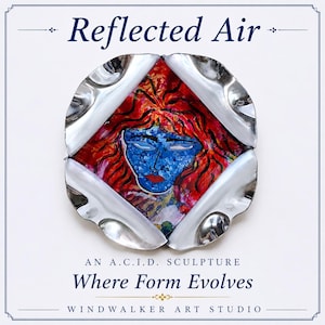May include: A circular metallic sculpture with a central diamond-shaped cutout revealing a painted face with blue skin, red hair, and red lips. The text Reflected Air, An A.C.I.D. Sculpture, Where Form Evolves, and Windwalker Art Studio is visible.