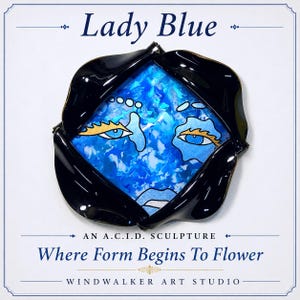 May include: A square-shaped sculpture titled Lady Blue, featuring a central blue and white marbled face with gold-accented eyes and lips, framed by four glossy black curved petals. Text reads Lady Blue, An A.C.I.D. Sculpture, Where Form Begins To Flower, Windwalker Art Studio.