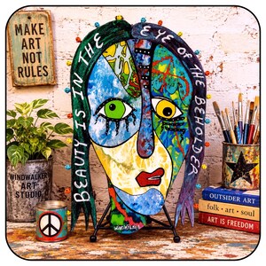May include: A colorful, abstract wooden face sculpture featuring the text "BEAUTY IS IN THE EYE OF THE BEHOLDER" painted along the hair. The face has one green eye and one yellow eye, red lips, and is supported by a black metal stand.