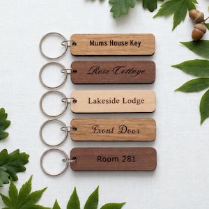 Könnte beinhalten: Fünf Holzschlüsselanhänger mit silbernen Schlüsselringen. Jeder Schlüsselanhänger ist rechteckig und mit Text graviert: "Mums House Key", "Rose Cottage", "Lakeside Lodge", "Front Door" und "Room 281". Die Schlüsselanhänger sind in verschiedenen Brauntönen gehalten.