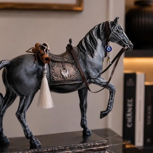 Puede incluir: Estatua de caballo gris oscuro con silla de montar y brida. La estatua tiene una silla decorativa con una borla blanca y un amuleto de ojo azul en la brida. El caballo está sobre una base rectangular con detalles ornamentados.