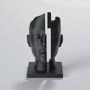 May include: A black sculpture of a human head split into two vertical sections. The head is mounted on a square base. The sculpture has a minimalist design and a matte finish, with closed eyes and a neutral expression.