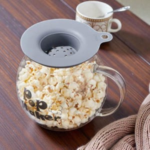 May include: A clear glass popcorn maker filled with popped popcorn, topped with a gray lid. The words "Pop Maker" are printed on the side. A white and brown mug with a spoon and a pink scarf are in the background.