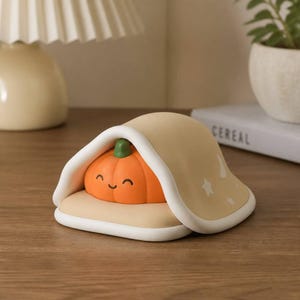 Puede incluir: Una pequeña figura de calabaza naranja con una cara sonriente que descansa bajo una cubierta de color beige con forma de manta. La cubierta presenta un borde blanco y un patrón de pequeñas estrellas y lunas crecientes blancas en el lateral.