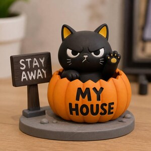 Pode incluir: Uma estatueta de gato preto com uma expressão zangada sentada dentro de uma abóbora laranja. A abóbora tem o texto MY HOUSE impresso. Uma placa castanha escura ao lado da abóbora diz STAY AWAY. O objeto assenta numa base circular cinzenta.