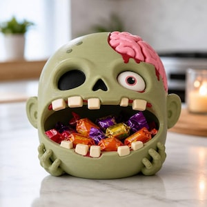 Könnte beinhalten: Eine hellgrüne Schale in Form eines Zombiekopfes mit weit geöffnetem Mund, gefüllt mit bunt verpackten Bonbons. Die Figur zeigt ein rosa freiliegendes Gehirn oben links, eine schwarze Augenhöhle, ein weißes Auge mit rosa Iris und cremefarbene Zähne.
