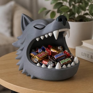 May include: A gray wolf head-shaped bowl with a wide-open mouth featuring sharp white teeth. The interior of the mouth is filled with various individually wrapped chocolate candy bars including Twix, Snickers, and Milky Way brands.