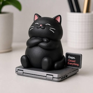 Könnte beinhalten: Eine mattschwarze Katzenfigur mit rosa Innenohren und lächelndem Gesichtsausdruck, die mit verschränkten Armen auf einem grauen Klappgerät sitzt. Ein kleines rechteckiges Schild an der Basis zeigt den weiß-roten Text: Oops. Closed Everything.