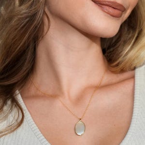 May include: A gold necklace with a pendant in the shape of an oval. The pendant is a light, pearly color. The woman in the image is wearing a cream-colored ribbed shirt.