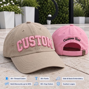 May include: A tan baseball cap with the word CUSTOM embroidered in pink block letters on the front, and a pink baseball cap with the words Custom Text embroidered in black script on the back, featuring an adjustable strap with a metal buckle.