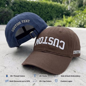 May include: A navy blue baseball cap with the text CUSTOM TEXT embroidered on the back and a brown baseball cap with the text CUSTOM embroidered in white on the front and side. Both caps feature adjustable metal buckles and structured fabric panels.