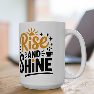 Puede incluir: Taza de cerámica blanca con las palabras "Rise and Shine" en letras negras y doradas. El diseño incluye un sol, estrellas y una taza de café. La taza está sobre una superficie de madera.