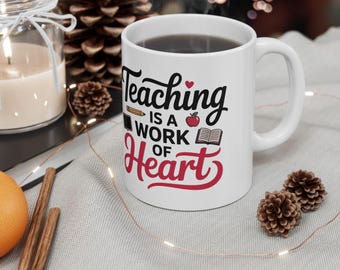 以下が含まれることがあります： 「Teaching is a Work of Heart」の文字が黒と赤でプリントされた白いセラミックマグ。マグには鉛筆、リンゴ、本のグラフィックが描かれています。マグには濃い色の液体が入っています。