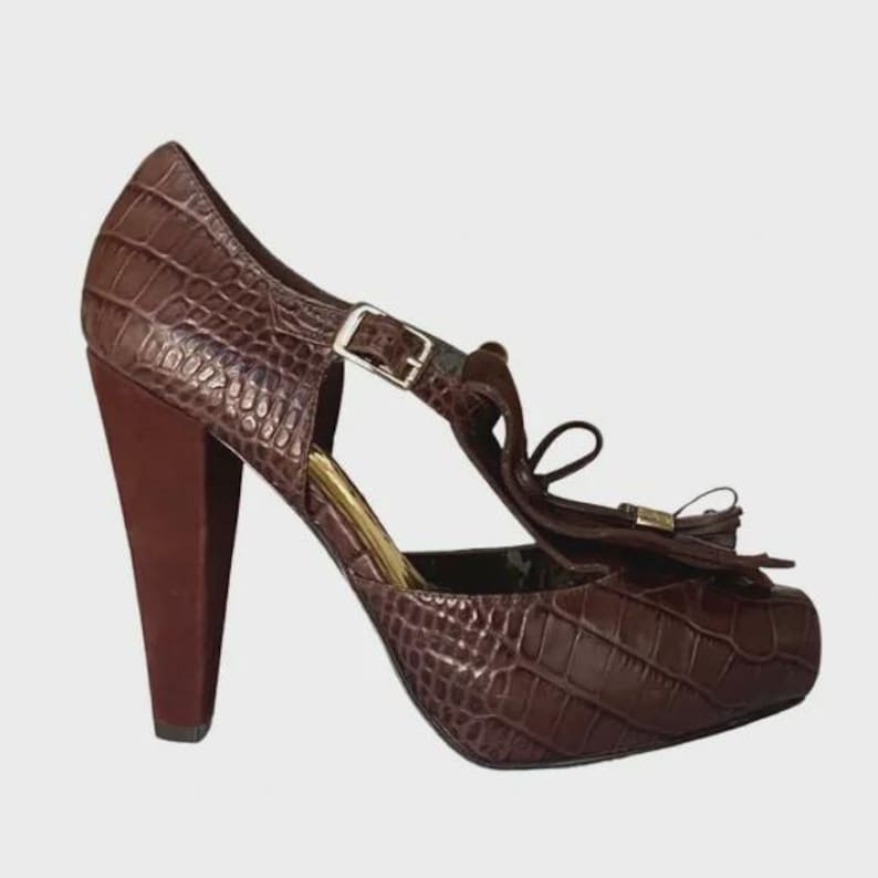 May include: Brown high-heeled shoe with a textured, crocodile-like pattern. The shoe features a T-strap with a buckle, a bow detail, and a thick heel. The heel is approximately 4 inches tall.