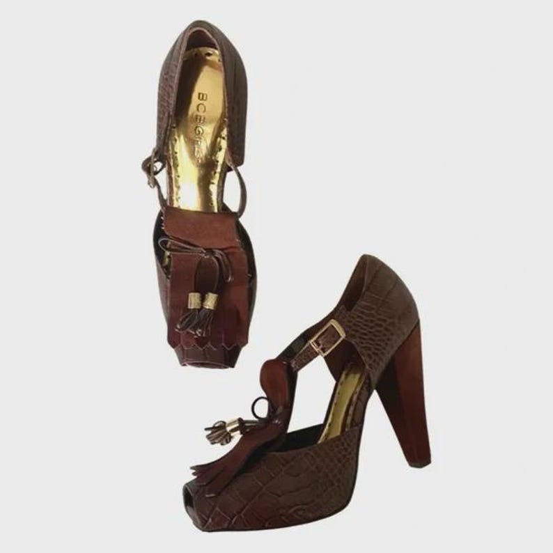 May include: Brown high-heeled shoes with a textured pattern and a peep-toe design. The shoes have a t-strap with a buckle closure and a tassel detail. The heels are a dark brown color. The interior of the shoe is gold. The brand name "BCBG" is visible.