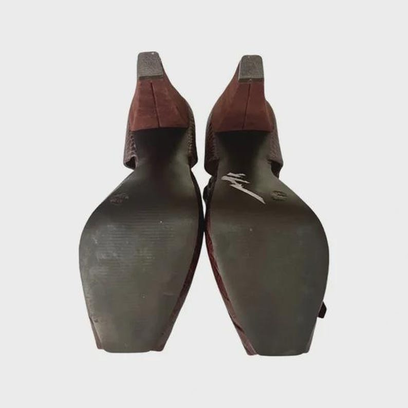 May include: Pair of burgundy-colored women's heels with a textured, snakeskin-like pattern. The shoes have a square toe and a block heel. The soles are dark brown. The heels are approximately 3 inches high.