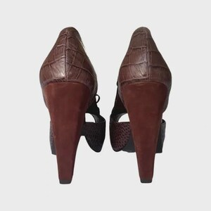 May include: A pair of brown high-heeled shoes. The heels are a dark brown, and the uppers have a textured, crocodile-like pattern. The shoes have a strap design and are set against a neutral background.