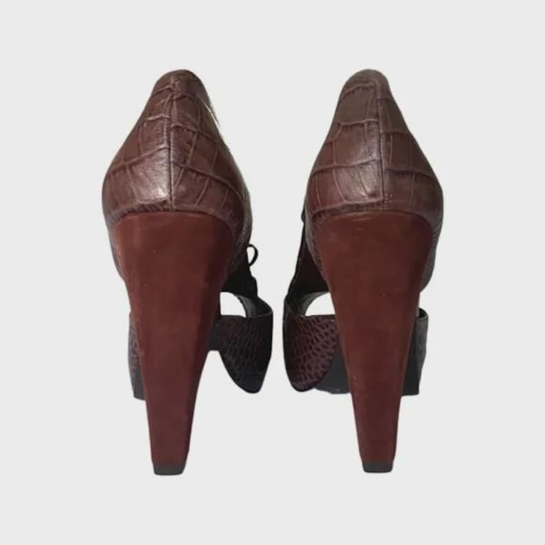May include: A pair of brown high-heeled shoes. The heels are a dark brown, and the uppers have a textured, crocodile-like pattern. The shoes have a strap design and are set against a neutral background.