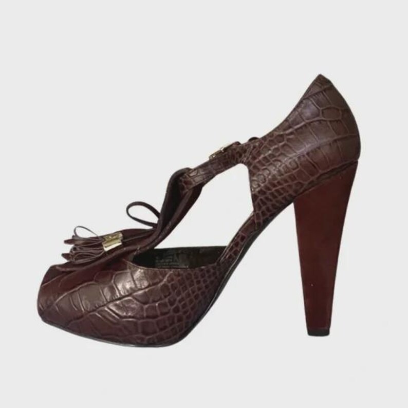 May include: Brown platform heels with a faux crocodile skin texture. The heels have a rounded toe, a strap across the top of the foot, and a high, dark brown heel. A small tassel and gold-colored hardware add detail.