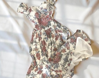 May include: A vintage-style dress with a floral pattern in shades of cream, burgundy, and blue. The dress features a fitted bodice, off-the-shoulder sleeves, and a flowing skirt. The fabric appears to be lightweight, suitable for warmer weather.
