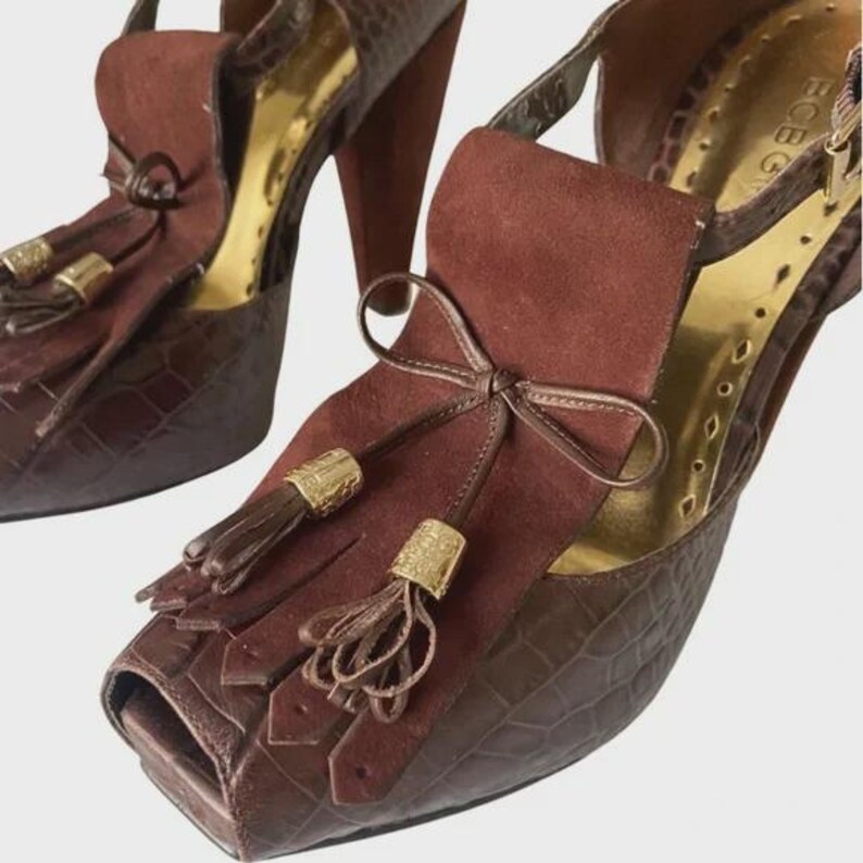May include: A pair of brown high-heeled shoes with a peep-toe design. The shoes feature a crocodile-textured pattern on the base and a suede-like material on the upper, adorned with tassels and a bow. The heels are approximately 4 inches tall.