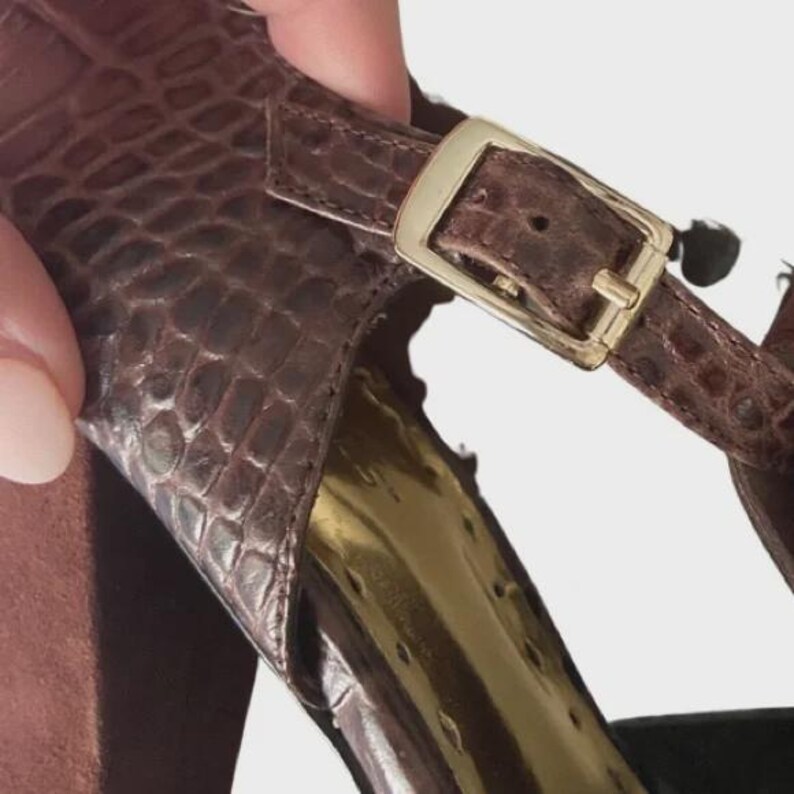 May include: Close-up of a brown high-heeled sandal with a textured, reptile-like pattern. The sandal features a gold-colored buckle and a metallic gold interior. The heel is a solid brown color.