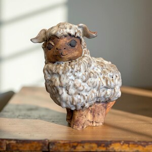 May include: A ceramic sheep figurine with a textured, off-white woolly coat and a brown face. The sheep has small, curved horns and is standing on a brown base. The figurine is a decorative item.