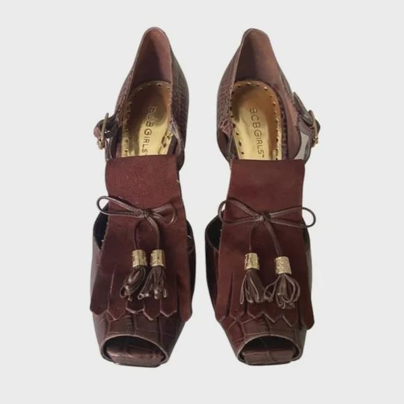 May include: A pair of brown open-toe heels with a textured, crocodile-like pattern. The heels feature a suede-like flap with tassels and a bow detail. The brand name "BCBG GIRLS" is visible on the insole.