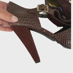 May include: Brown faux-crocodile leather sandal with a T-strap design and a gold-colored buckle. The sandal features a dark brown, cone-shaped heel. The shoe is held up by a hand.