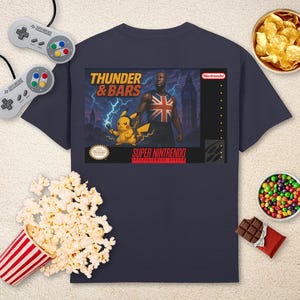 May include: Navy blue t-shirt with a graphic inspired by a Super Nintendo game. The graphic includes the text "Thunder & Bars", a man wearing a Union Jack, and Pikachu. The shirt is surrounded by popcorn, chips, candy, and game controllers.