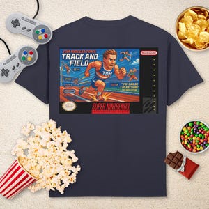 Puede incluir: Camiseta azul marino con un diseño gráfico de videojuego retro. El gráfico dice "Tom Hiddleston's Track and Field" con una ilustración de corredor. La imagen incluye snacks y un mando de videojuego retro.
