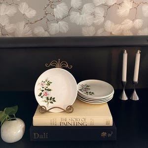 May include: White ceramic plates with a floral rose design, displayed on a gold-colored stand. A stack of matching plates is visible. The plates are set on top of books. Two silver candle holders with white candles are on the right.